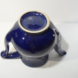 May include: A cobalt blue ceramic teapot with a unique three-legged design. The teapot has a round body and a spout on one side. The bottom of the teapot has the words "MADE IN CANADA" printed on it.