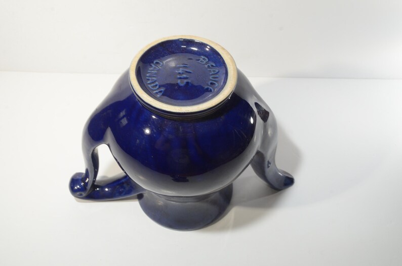 May include: A cobalt blue ceramic teapot with a unique three-legged design. The teapot has a round body and a spout on one side. The bottom of the teapot has the words "MADE IN CANADA" printed on it.