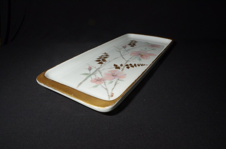 May include: Rectangular white serving tray with a gold rim and floral design. The tray features pink flowers and gold accents on a white background. A decorative piece for serving or display.