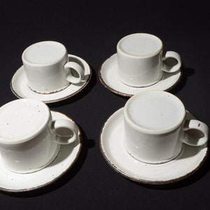 May include: Four white ceramic coffee cups with brown rims and speckles on the surface, each sitting on a matching saucer.