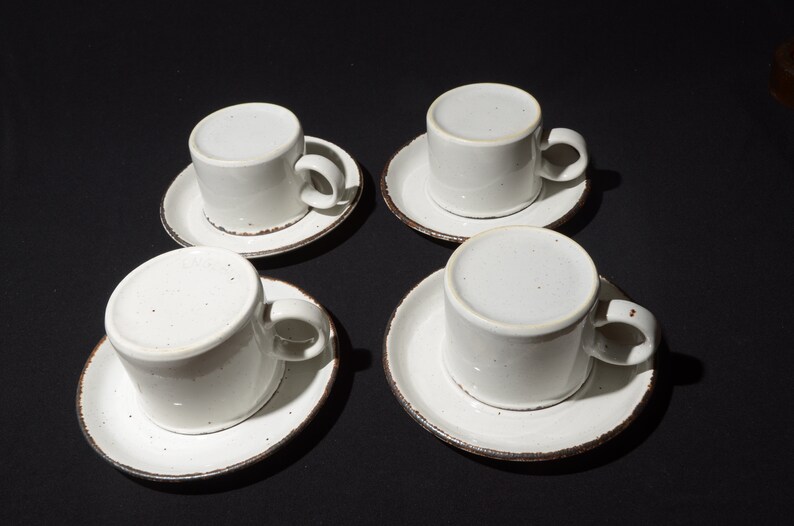 May include: Four white ceramic coffee cups with brown rims and speckles on the surface, each sitting on a matching saucer.
