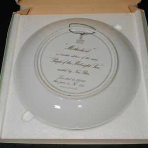 May include: A white decorative plate with the text "Motherhood" and "People of the Midnight Sun" in a box. The plate has a silver rim and a wire for hanging. The plate is a limited edition.