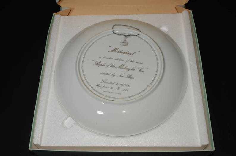 May include: A white decorative plate with the text "Motherhood" and "People of the Midnight Sun" in a box. The plate has a silver rim and a wire for hanging. The plate is a limited edition.