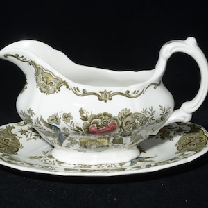May include: A white porcelain gravy boat and saucer set. The boat features a curved handle and spout, adorned with floral and foliage designs in green, brown, and red. The edges of the boat and saucer are scalloped. The set is suitable for serving gravy.