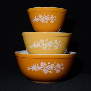 May include: Three vintage mixing bowls stacked, in shades of orange and yellow. Each bowl features a white floral design. The bowls are nested, with the largest at the bottom and the smallest at the top, against a black background.