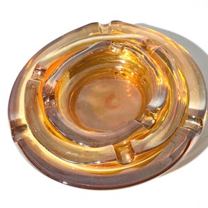 May include: A round, amber-colored glass ashtray featuring a nested design with a smaller circular basin centered within a larger circular base. The glass has a translucent, warm orange hue with smooth, curved edges and integrated notches.