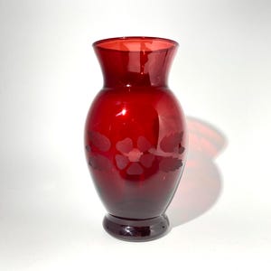 May include: A translucent ruby red glass vase with a bulbous body and a flared rim. The exterior surface features a frosted, etched floral pattern consisting of a central five-petaled flower flanked by two leaves on each side, set on a circular base.