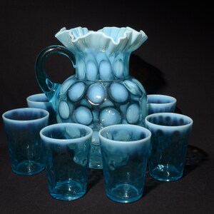 May include: A set of seven blue glass pieces, including a jug and six glasses. The jug has a ruffled top and a pattern of white circles. The glasses are all the same size and have a similar pattern of white circles.