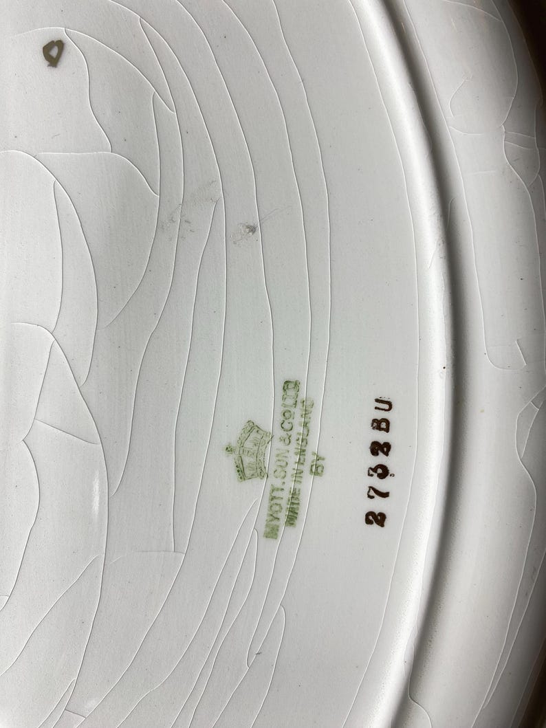 May include: Close-up of a white ceramic plate with a cracked glaze. The plate has a slightly raised edge. The words "MYOTT, SON & CO LTD MADE IN ENGLAND" are printed in green, along with the number "2732 BU".