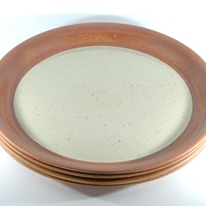 May include: A stack of three round ceramic plates featuring a wide, smooth terracotta-colored rim and a speckled, off-white center. The plates have a consistent circular design with a raised edge and a matte finish on the outer rim.