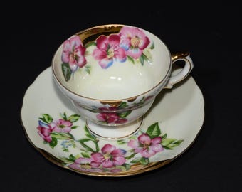 以下が含まれることがあります： クリーム色の磁器製ティーカップとソーサーのセット。ピンクの花と緑の葉が手描きで描かれています。カップの縁とソーサーのスカラップ状の縁には金色のトリムが施されており、カップには持ち手が付いています。