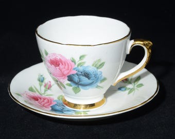 May include: White teacup and saucer set with pink and blue floral design. The cup and saucer have gold trim. The cup handle and base are also gold. The floral design includes pink and blue roses with green leaves.