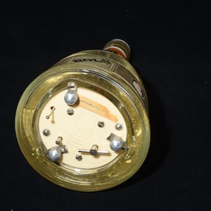 May include: A clear glass bottle with a gold-colored label and a cream-colored base. The base has three silver-colored metal feet and a small label. The bottle is lying on its side against a black background.