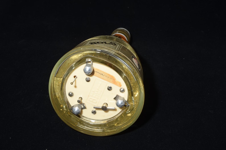 May include: A clear glass bottle with a gold-colored label and a cream-colored base. The base has three silver-colored metal feet and a small label. The bottle is lying on its side against a black background.