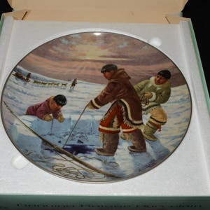 May include: A ceramic plate with a colorful painting of three people ice fishing on a frozen lake. The scene is set against a snowy background with a bright blue sky. The plate is in a white box with the words "Canadian Heritage Collection" printed on the bottom.