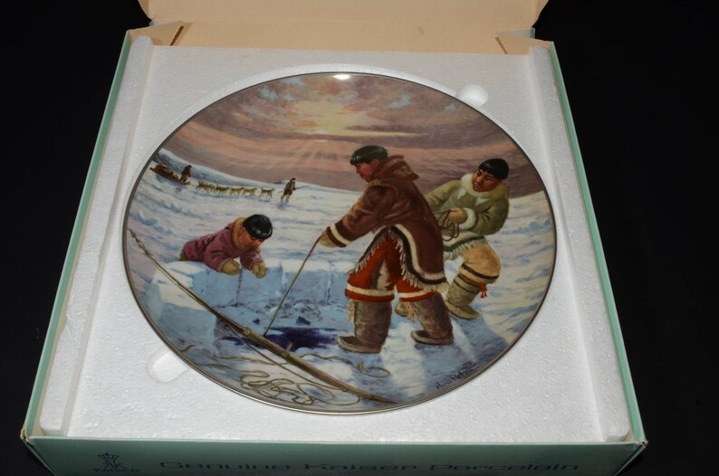 May include: A ceramic plate with a colorful painting of three people ice fishing on a frozen lake. The scene is set against a snowy background with a bright blue sky. The plate is in a white box with the words "Canadian Heritage Collection" printed on the bottom.
