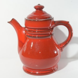 Puede incluir: Una tetera de cerámica roja con tapa y asa. La tetera tiene una banda marrón alrededor del medio.