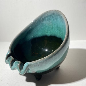 Puede incluir: Un cenicero de cerámica de color verde azulado y negro con un borde ondulado y tres pequeñas patas. El cenicero está esmaltado con un acabado moteado.