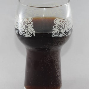 May include: A clear glass with a white Pepsi logo etched on the side, filled with a dark brown liquid. The glass has a wide base and a narrow top.