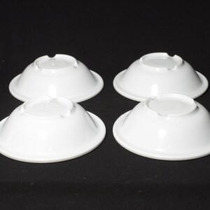 May include: Four white ceramic bowls, each with a slightly rounded bottom and a smooth, glossy finish. The bowls are arranged in a square pattern on a black surface.
