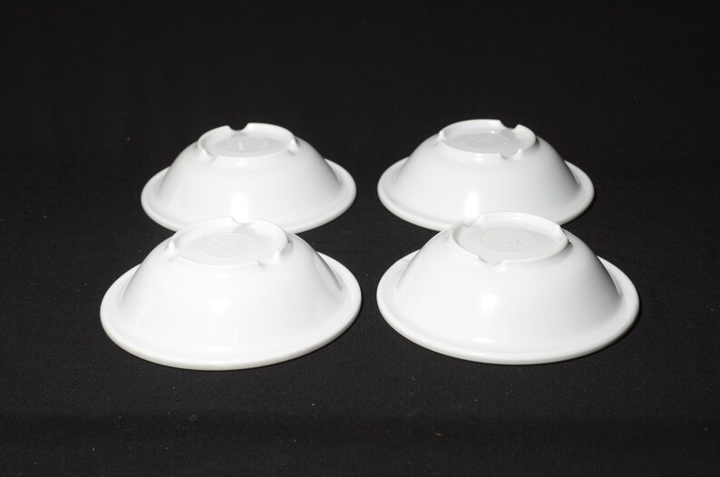 May include: Four white ceramic bowls, each with a slightly rounded bottom and a smooth, glossy finish. The bowls are arranged in a square pattern on a black surface.
