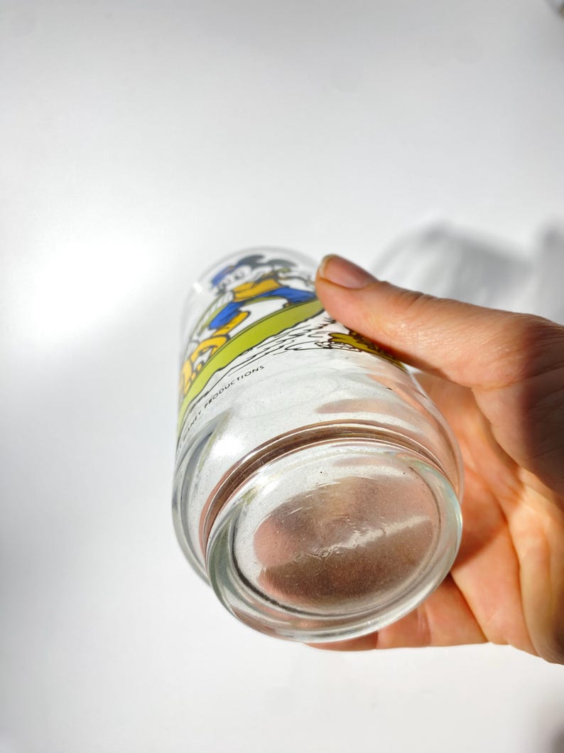 May include: A clear glass tumbler featuring a cartoon image of Donald Duck in his signature blue sailor suit. The glass has a wide base and a slightly tapered body. The image is in bright colors, with the words "Walt Disney Productions" printed on the glass.