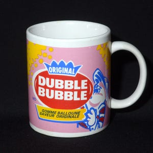 May include: A white ceramic mug with a pink background and a cartoon character. The mug features the text "Original Dubble Bubble Gomme Balloune Saveur Originale".