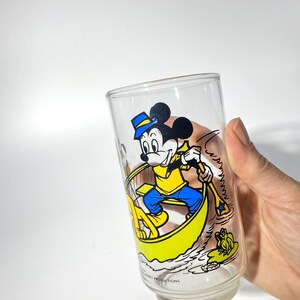 May include: Clear glass featuring Mickey Mouse in a yellow boat, wearing a blue hat and shirt, rowing with a paddle. The design includes a frog and water details. A vintage Disney collectible.