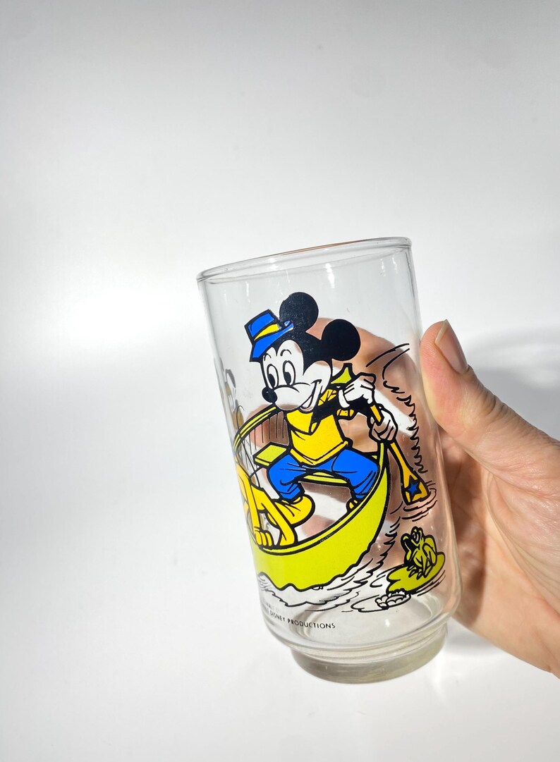 May include: Clear glass featuring Mickey Mouse in a yellow boat, wearing a blue hat and shirt, rowing with a paddle. The design includes a frog and water details. A vintage Disney collectible.