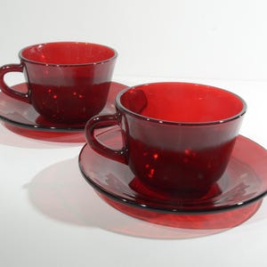 May include: Two red glass teacups and saucers. The cups and saucers are a deep, rich red colour. The cups have a rounded shape and a small handle. The saucers are slightly larger than the cups and have a flat bottom.