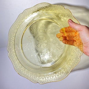 May include: A yellow glass plate with a scalloped edge and an intricate etched design. The plate has a gold rim.