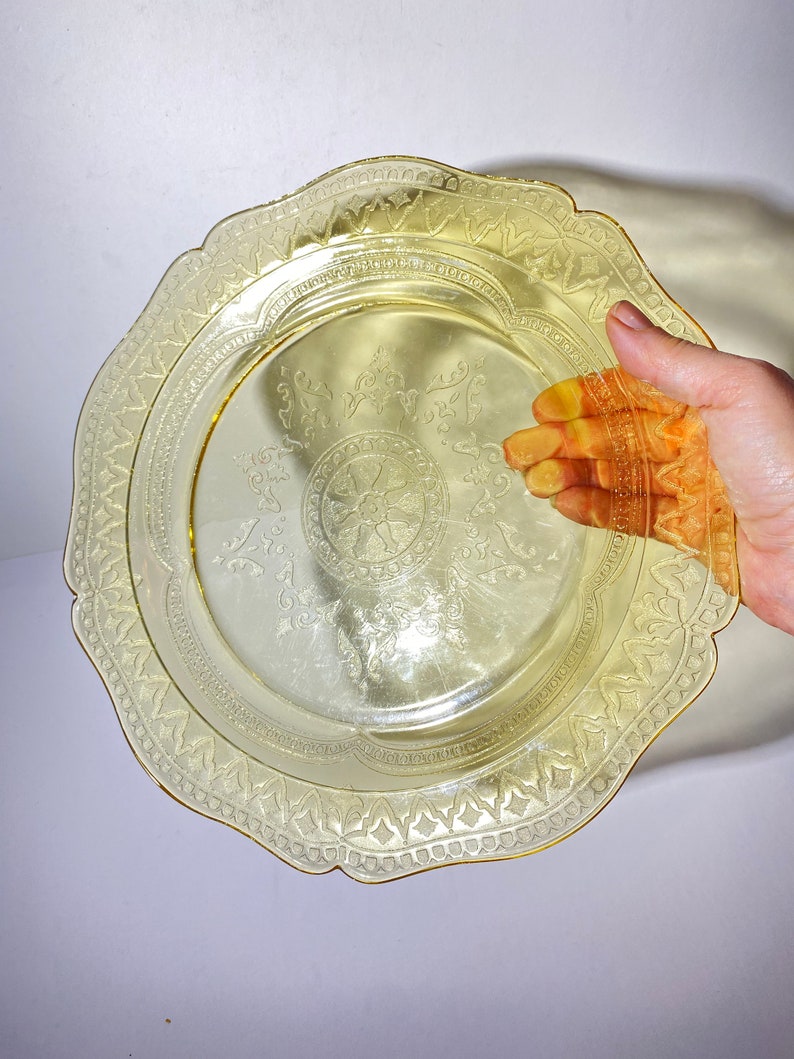 May include: A yellow glass plate with a scalloped edge and an intricate etched design. The plate has a gold rim.