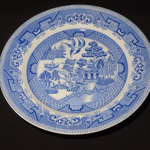 Op de afbeelding: Een blauw en wit keramisch dinerbord met een traditioneel wilgenmotief. Het bord heeft een centraal beeld van een pagode, een brug en een wilgenboom.