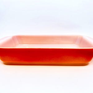 Puede incluir: Una fuente de horno rectangular con un acabado liso y brillante. El exterior presenta un color naranja vibrante y saturado que se transforma en un tono rosa pálido y suave en el interior y a lo largo del borde superior. Incluye asas laterales.