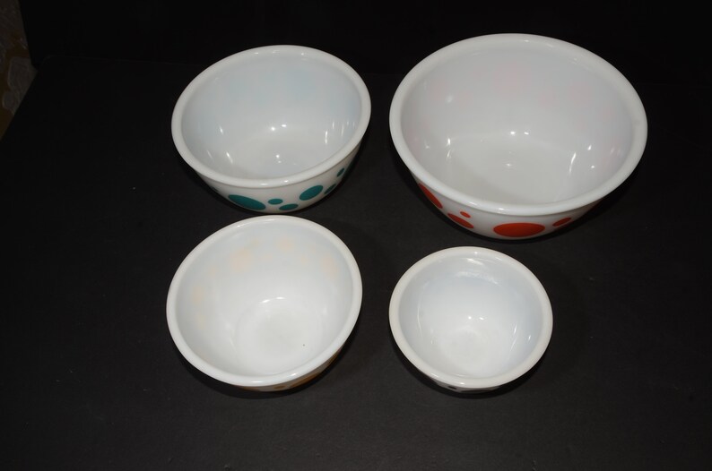 May include: Set of four white mixing bowls with colorful polka dot designs. The bowls are nested in size order, with the largest bowl having orange dots, the second largest bowl having teal dots, and the two smallest bowls having orange and yellow dots.
