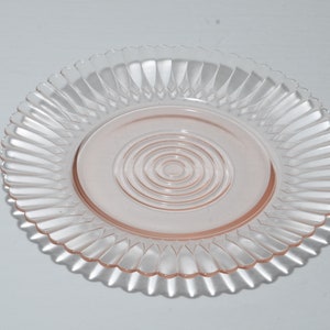 Puede incluir: Un plato de vidrio transparente te&ntilde;ido de rosa, con un borde acanalado y un dise&ntilde;o circular central. El dise&ntilde;o del plato presenta c&iacute;rculos conc&eacute;ntricos en el centro. El plato es probablemente para servir o exhibir.