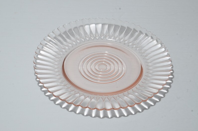 Puede incluir: Un plato de vidrio transparente te&ntilde;ido de rosa, con un borde acanalado y un dise&ntilde;o circular central. El dise&ntilde;o del plato presenta c&iacute;rculos conc&eacute;ntricos en el centro. El plato es probablemente para servir o exhibir.