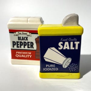 May include: Two ceramic salt and pepper shakers. The black pepper shaker is white with a red border and the words "Black Pepper" in large letters. The salt shaker is navy blue with yellow trim and the word "Salt".