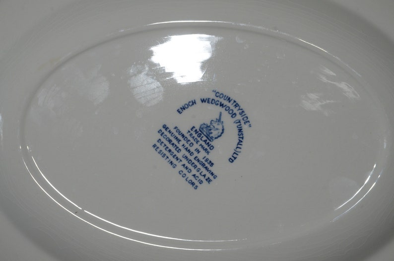 Op de afbeelding: Een wit ovaal bord met een blauw-wit logo met de tekst "COUNTRYSIDE" "ENOCH WEDGWOOD (TUNSTALL) LTD" "ENGLAND" "FOUNDED IN 1835" "TRADE MARK" "GENUINE HAND ENGRAVING" "DECORATED UNDERGLAZE" "DETERGENT AND ACID RESISTING COLORS". Het logo bevat ook een eenhoorn.