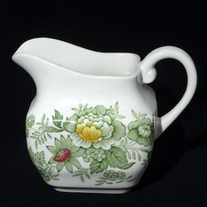 Pode incluir: Leiteira de cerâmica branca com um design floral. A leiteira apresenta um bico curvo e uma pega. O padrão floral inclui folhas verdes, flores amarelas e brancas e detalhes vermelhos. A leiteira está sobre um fundo preto.