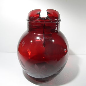 May include: A red glass pitcher with a wide mouth and a handle. The pitcher has an etched design of leaves on the side.