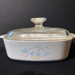 K&ouml;nnte beinhalten: Wei&szlig;e Auflaufform mit Glasdeckel und einem Blumendesign mit blauen Blumen und einem Korb mit Schleife. Das Design ist auf wei&szlig;em Hintergrund mit blauen Punkten.