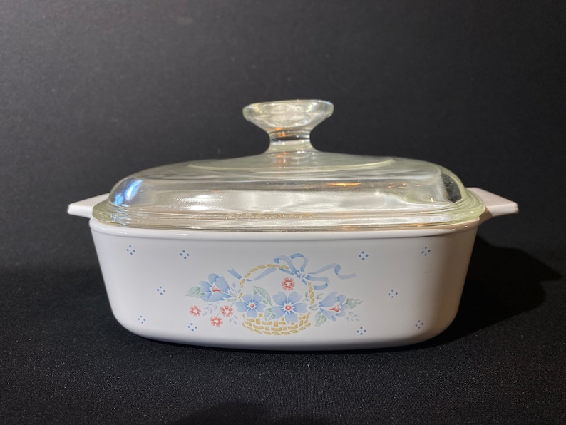 K&ouml;nnte beinhalten: Wei&szlig;e Auflaufform mit Glasdeckel und einem Blumendesign mit blauen Blumen und einem Korb mit Schleife. Das Design ist auf wei&szlig;em Hintergrund mit blauen Punkten.
