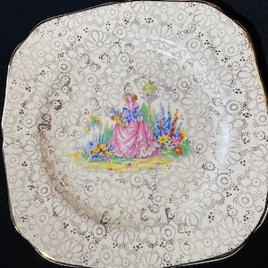 May include: A square, cream-coloured ceramic plate with a gold rim. The plate features a delicate lace pattern and a central image of a woman in a pink dress standing in a garden with flowers.