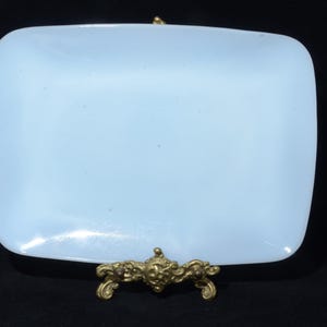 May include: A pale blue, rectangular dish with rounded corners, supported by an ornate, gold-coloured metal stand. The stand has a detailed design with a central face and scrollwork. The dish is smooth and appears to be made of glass or ceramic.