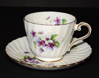 May include: A white teacup and saucer set with a floral pattern of purple and white flowers. The cup has a gold rim and the saucer has a scalloped edge.