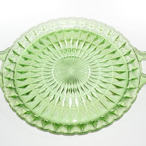 Puede incluir: Una bandeja redonda de vidrio verde claro con un patrón de diamantes y dos asas. La bandeja tiene un borde festoneado y es adecuada para servir alimentos o exhibir objetos decorativos. La bandeja tiene aproximadamente 30 cm de diámetro.