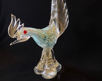 Könnte beinhalten: Eine Glasfigur eines Hahns mit gold- und grün schimmerndem Finish. Der Hahn hat ein rotes Auge und einen hochgestellten Schwanz.
