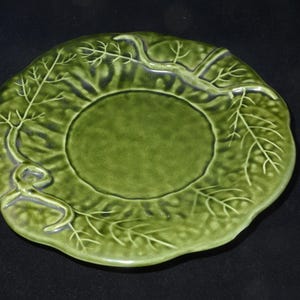 Può includere: Un piatto in ceramica verde con un design a foglie. Il piatto ha un centro rotondo e un bordo smerlato con dettagli di foglie e rami in rilievo. Il piatto è di colore verde pieno.