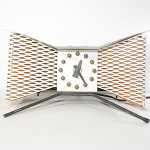 May include: A white and brown metal clock with a geometric design. The clock has a square face with black hands and gold accents. The clock is on a black metal stand with three legs.